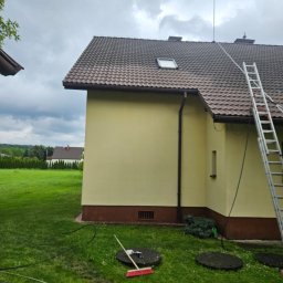 PROCLEAN360 GRZEGORZ PIKUŁA - Żółty dom z brązowym dachem i drabiną opartą o ścianę. Na trawniku widoczne narzędzia i włazy kanalizacyjne. Pochmurne niebo w tle.