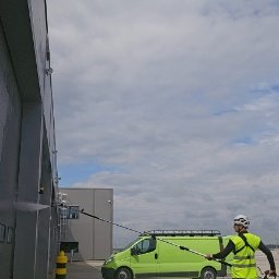 PROCLEAN360 GRZEGORZ PIKUŁA - Pracownik w kasku i kamizelce odblaskowej czyści elewację budynku przemysłowego za pomocą myjki ciśnieniowej. W tle zielony bus i fragment parkingu.