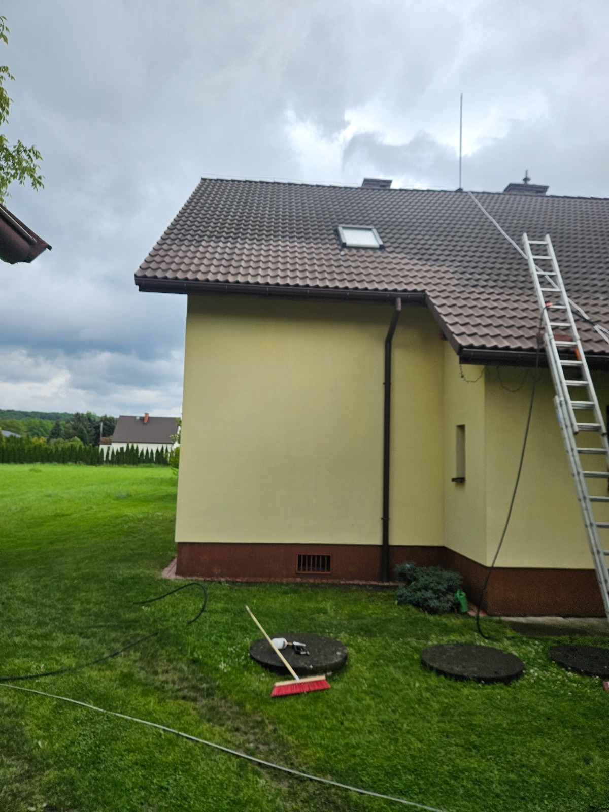 Żółty dom z brązowym dachem i drabiną opartą o ścianę. Na trawniku widoczne narzędzia i włazy kanalizacyjne. Pochmurne niebo w tle.