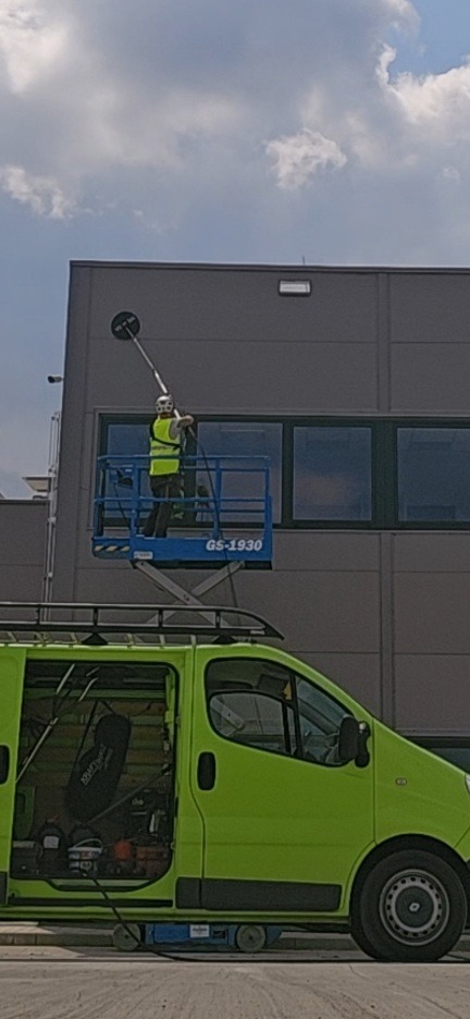 Pracownik w żółtej kamizelce, na podnośniku GS-1930, czyści elewację budynku przemysłowego za pomocą specjalistycznej myjki ciśnieniowej. Zielony bus z otwartymi drzwiami w tle.
