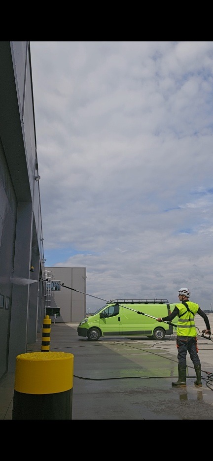 Pracownik w kasku i kamizelce myje szarą elewację budynku przemysłowego myjką ciśnieniową. W tle zielony bus i błękitne niebo.