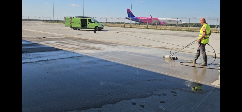 Pracownik w kamizelce odblaskowej czyści posadzkę przemysłową za pomocą myjki ciśnieniowej. W tle zielony bus i samolot na płycie lotniska.