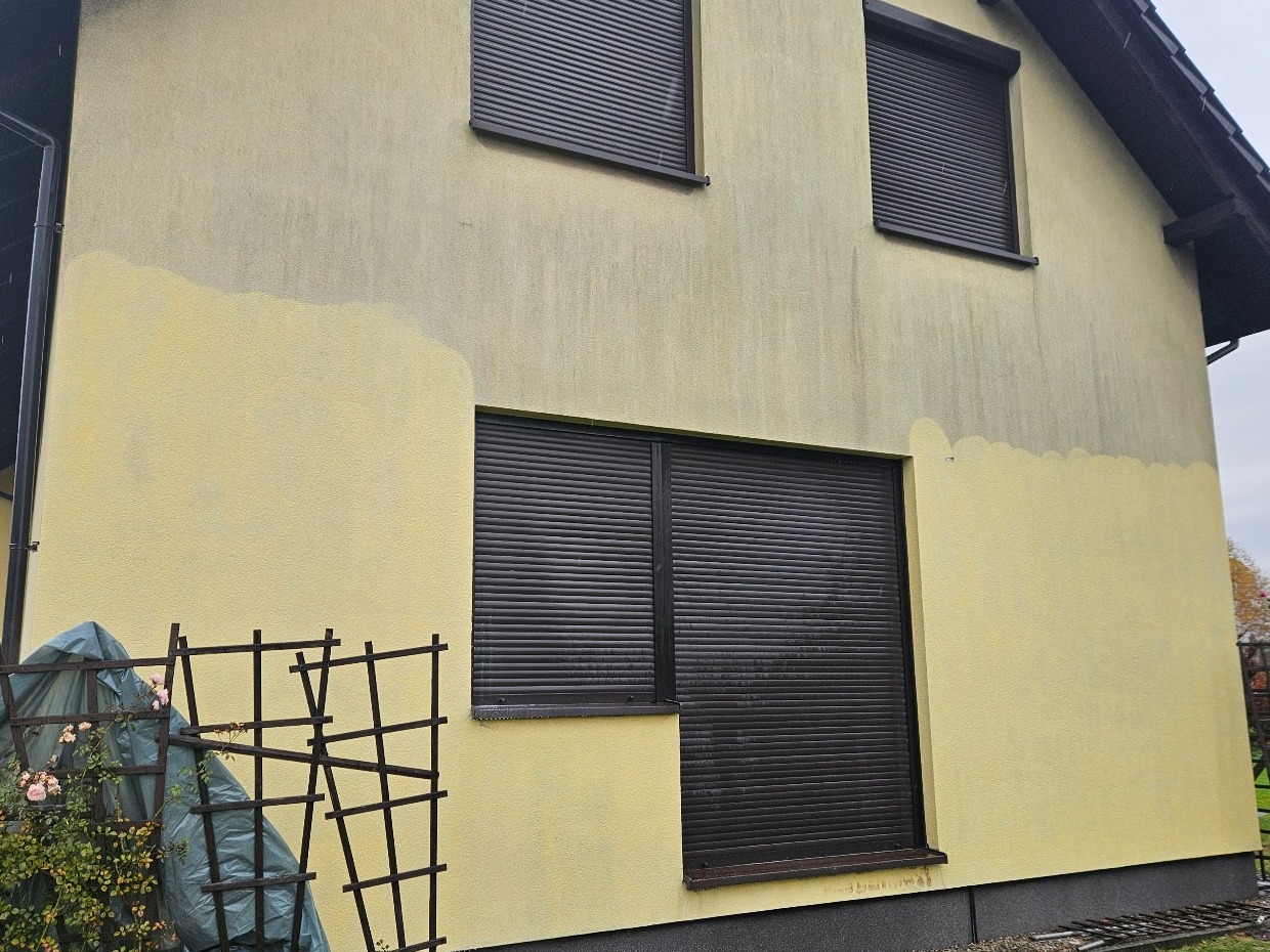 Żółta elewacja domu z brązowymi roletami, częściowo umyta, ukazująca różnicę w kolorze. Widoczny fragment ogrodu z roślinami i metalową konstrukcją.