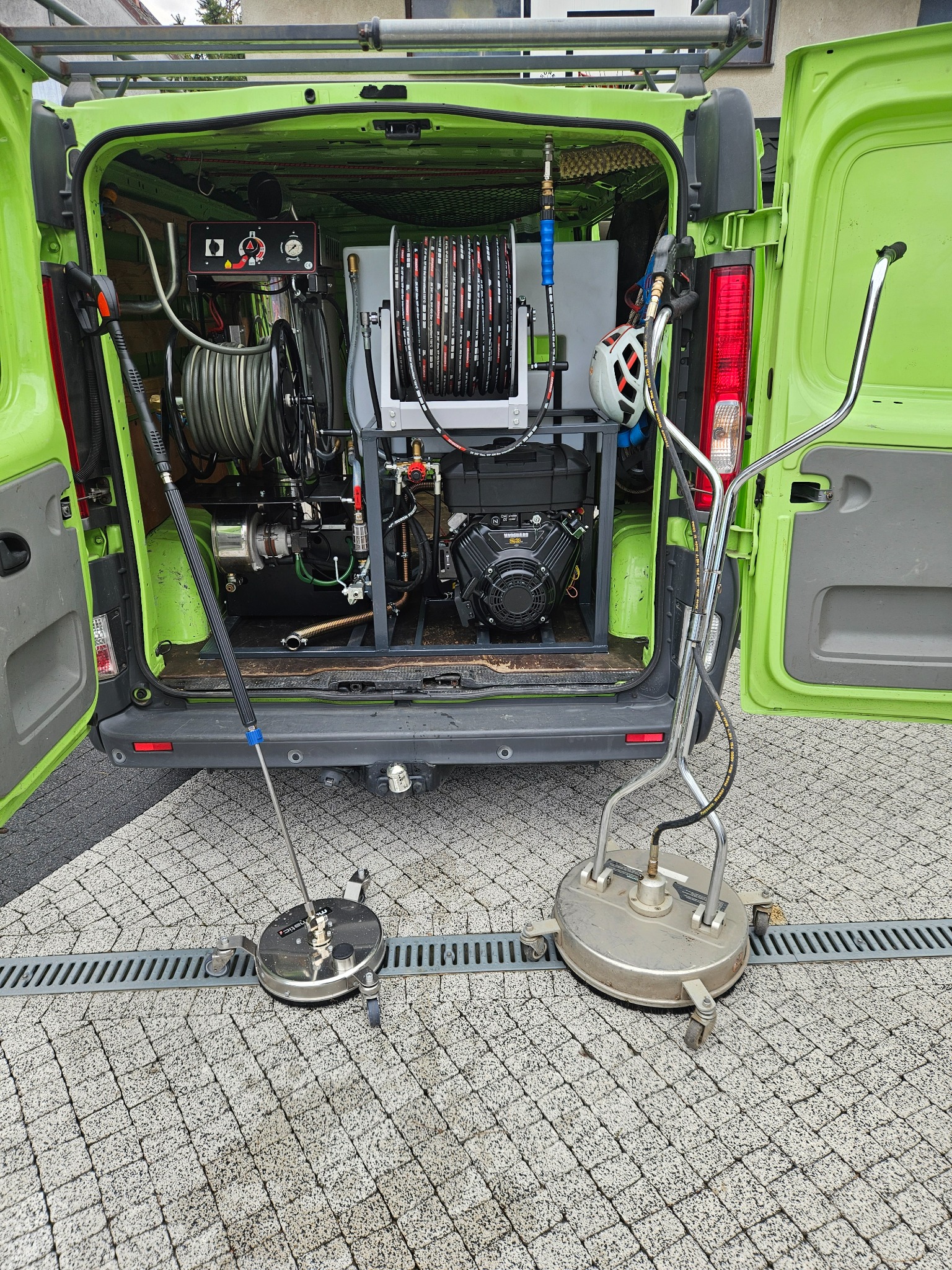Wnętrze zielonego busa z profesjonalnym sprzętem do czyszczenia ciśnieniowego. Na zewnątrz dwie okrągłe dysze do mycia kostki brukowej na szarej kostce.