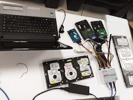 Stół z laptopem i kilkoma dyskami twardymi podłączonymi do specjalistycznego urządzenia do diagnostyki i odzyskiwania danych. Widoczne kable i zasilacze.