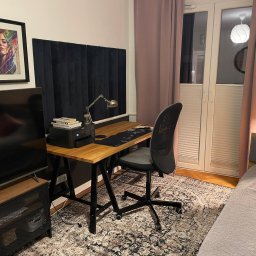 M.T Studio Design - Pokój z biurkiem z blatem z jasnego drewna i czarnymi nogami, krzesłem biurowym, lampką i zasłonami w odcieniach różu. Na ścianie obraz w czarnej ramie.