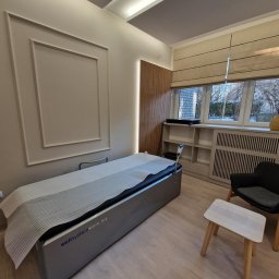 M.T Studio Design - Jasne wnętrze gabinetu z łóżkiem Wellsystem Medical Plus, krzesłem i stolikiem. Dekoracyjne panele ścienne i okno z żaluzjami rzymskimi.