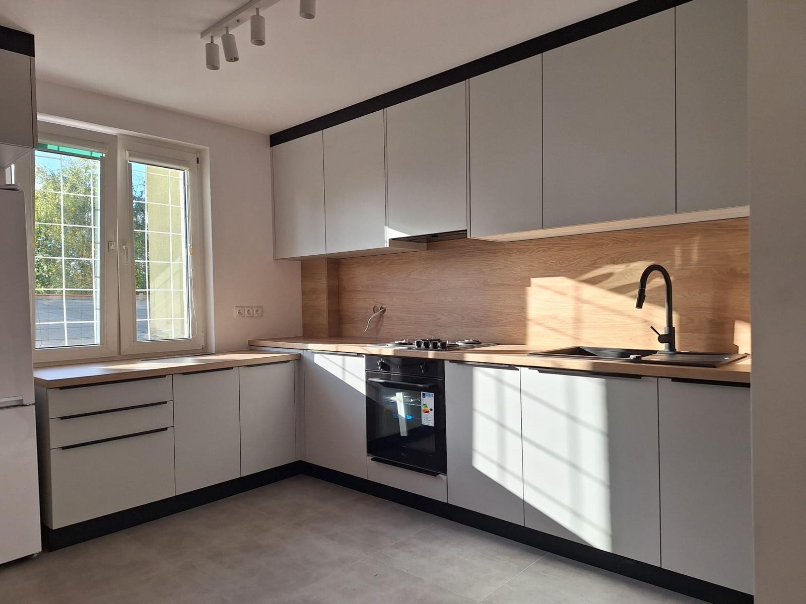 Jasna kuchnia z drewnianym blatem i czarnymi akcentami, oświetlona promieniami słońca wpadającymi przez okno. Nowoczesny minimalizm w aranżacji wnętrza.