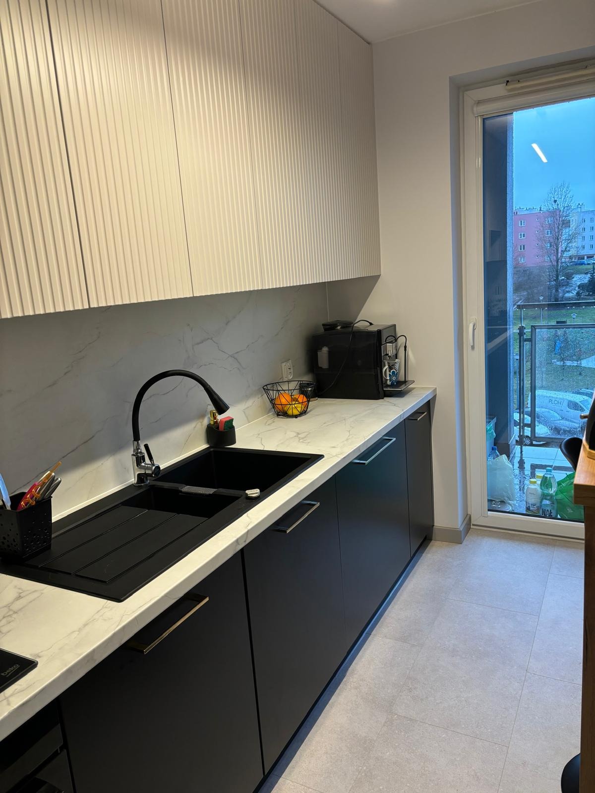 Nowoczesna kuchnia: czarne szafki z marmurowym blatem i ryflowane białe szafki wiszące. Czarny zlew i bateria. Widok na balkon przez okno.
