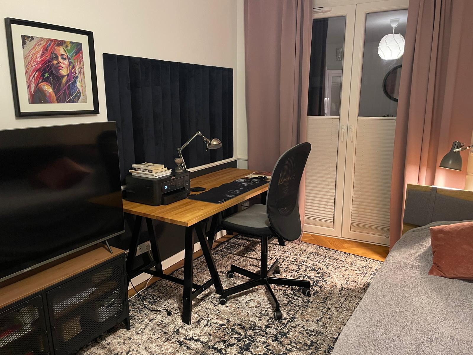 Pokój z biurkiem z blatem z jasnego drewna i czarnymi nogami, krzesłem biurowym, lampką i zasłonami w odcieniach różu. Na ścianie obraz w czarnej ramie.