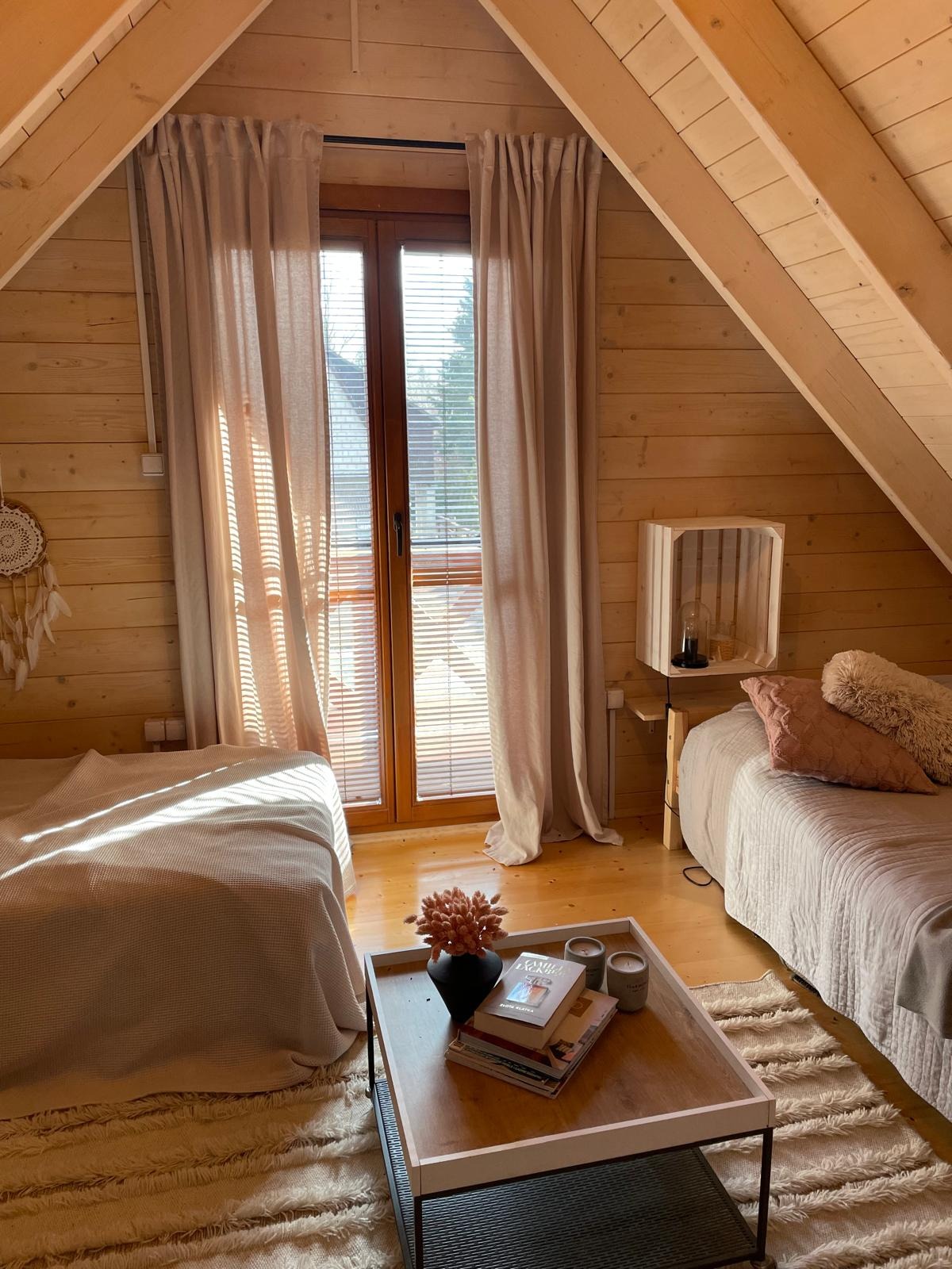 Przytulna aranżacja poddasza z drewnianymi ścianami. Dwa łóżka, stolik kawowy z książkami i kwiatami, okno z żaluzjami i zasłonami w ciepłych, naturalnych barwach.