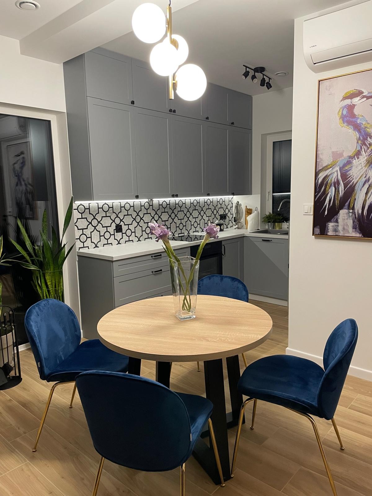 Szare szafki kuchenne z geometrycznym backsplashiem, okrągły stół z niebieskimi krzesłami i kwiatami w wazonie. Nowoczesne oświetlenie i drewniana podłoga.