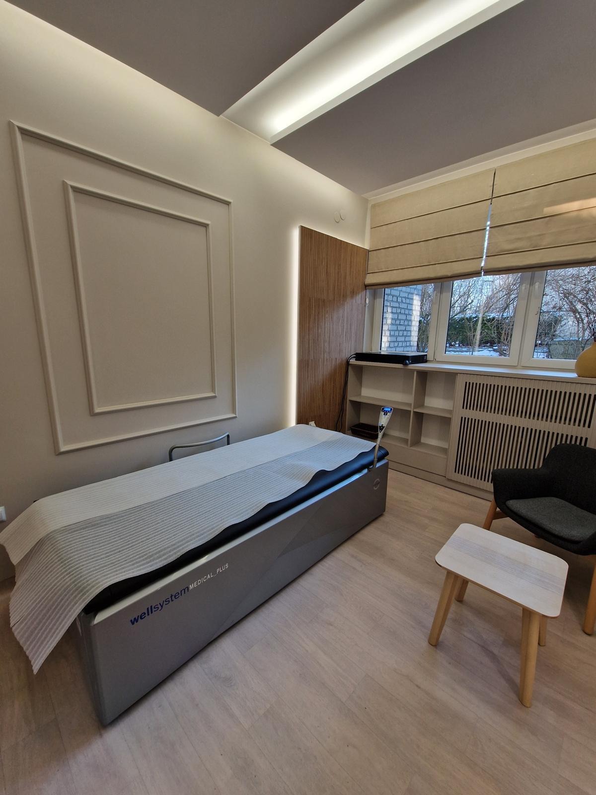 Jasne wnętrze gabinetu z łóżkiem Wellsystem Medical Plus, krzesłem i stolikiem. Dekoracyjne panele ścienne i okno z żaluzjami rzymskimi.
