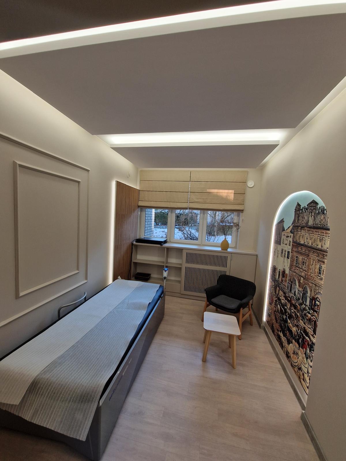 Jasne wnętrze gabinetu z łóżkiem do masażu, krzesłem i dekoracyjną fototapetą w kształcie łuku. Subtelne oświetlenie LED podkreśla nowoczesny charakter.