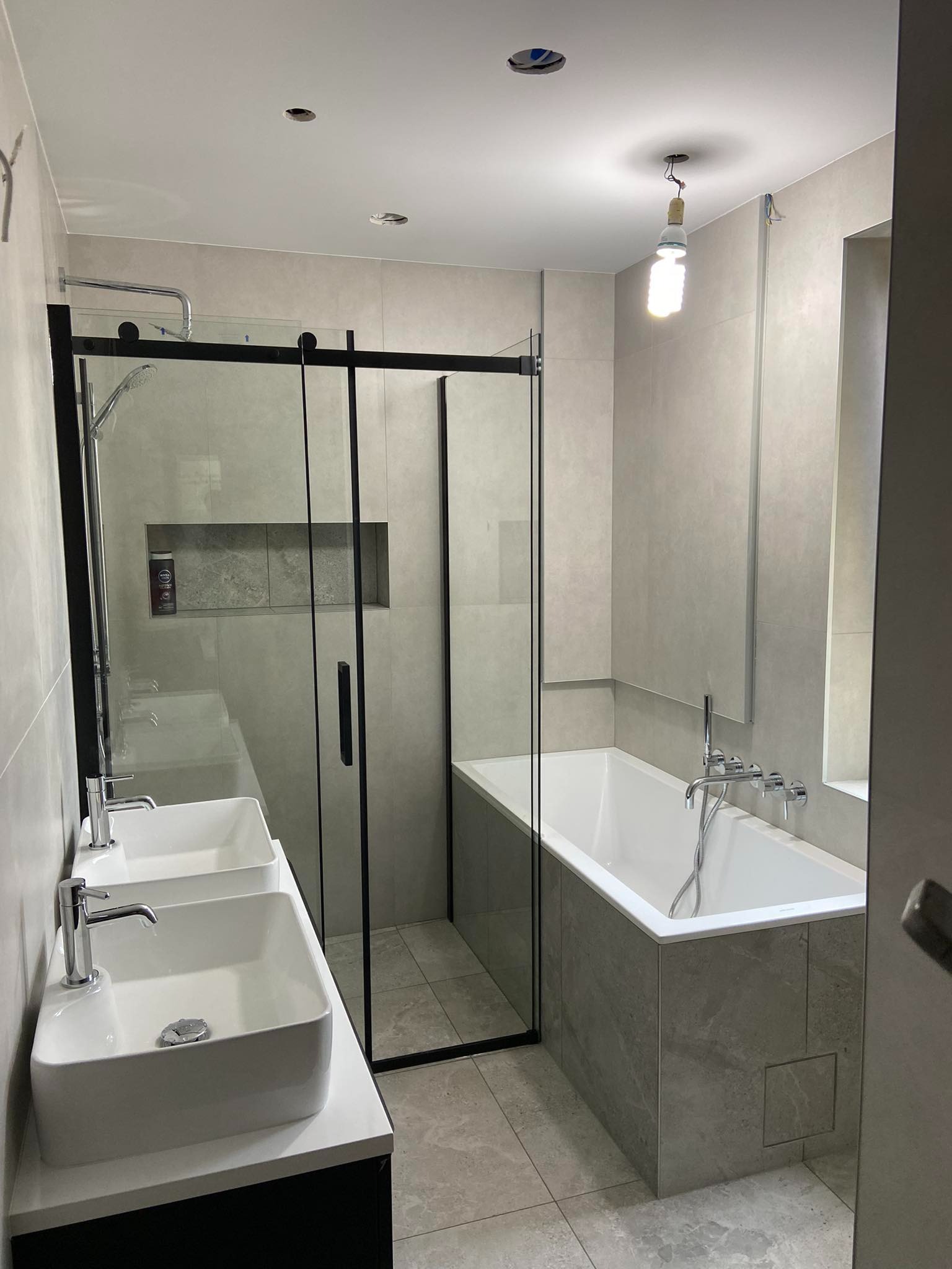 Nowoczesna łazienka z dwoma białymi umywalkami, kabiną prysznicową z czarnymi ramami i wanną w szarych płytkach. Widoczne oświetlenie tymczasowe.