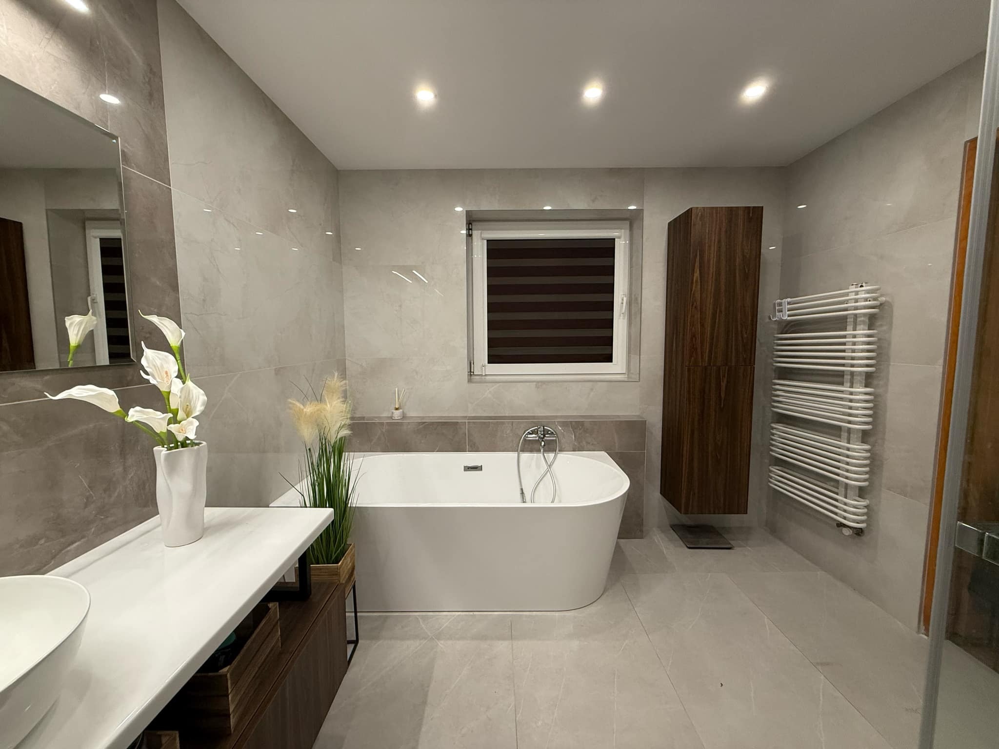Elegancka łazienka w odcieniach szarości z wolnostojącą wanną, umywalką na blacie i grzejnikiem drabinkowym. Minimalistyczny wystrój z akcentami roślinnymi.