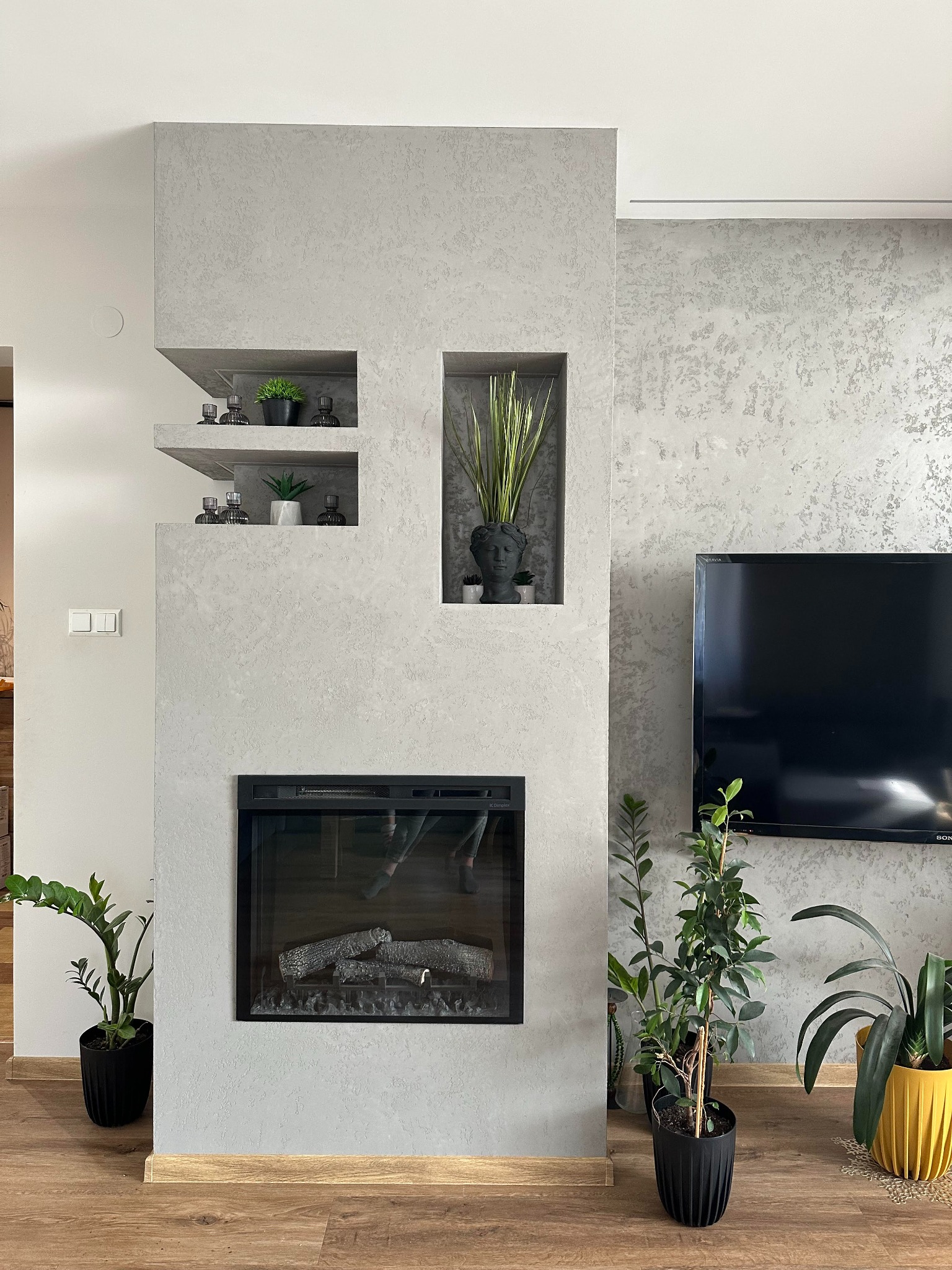 Szara ściana z wnękami, w których umieszczono rośliny doniczkowe i dekoracje. Poniżej wnęka z imitacją kominka. Na ścianie wisi czarny telewizor.