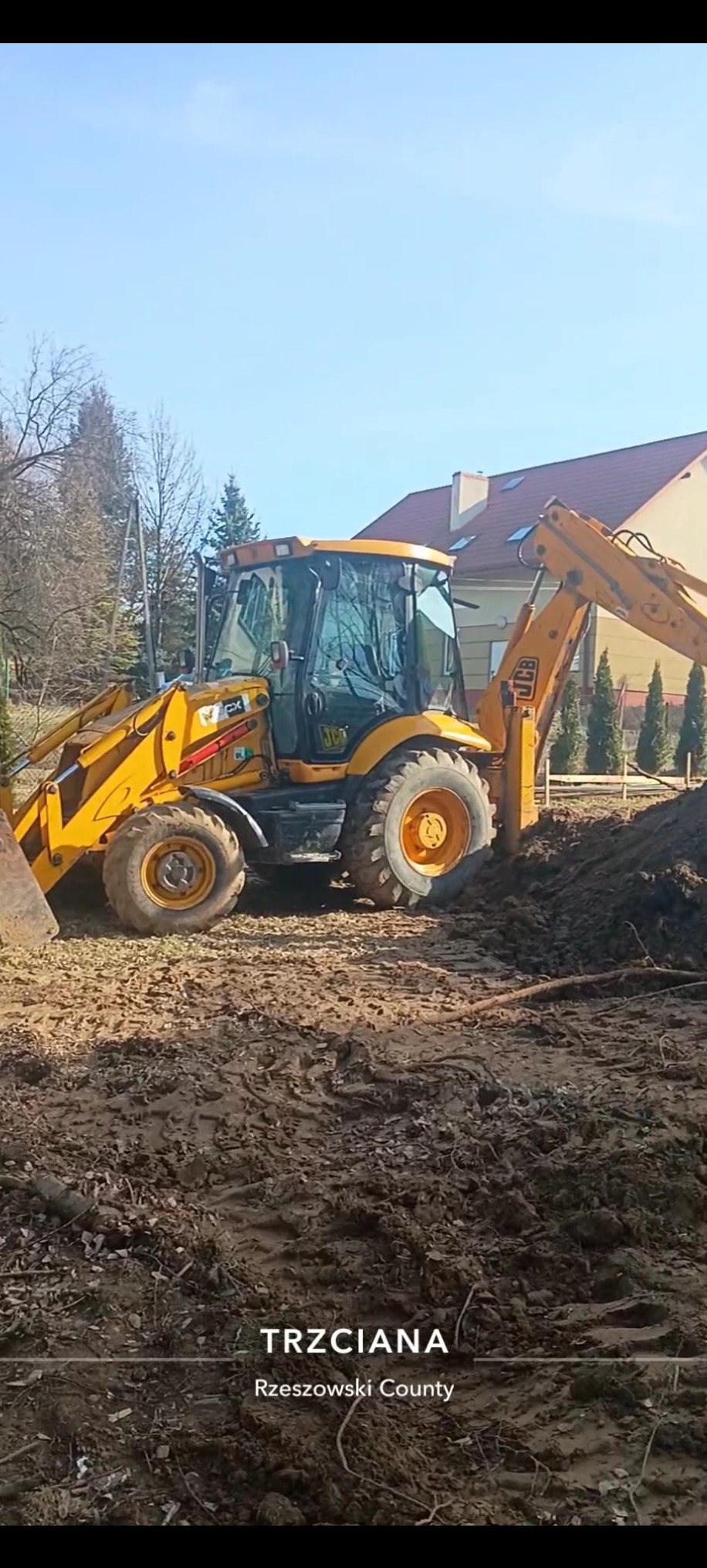Żółta koparko-ładowarka JCB na placu budowy, w tle dom i drzewa. Maszyna przygotowuje teren pod budowę, widoczna świeżo przekopana ziemia.