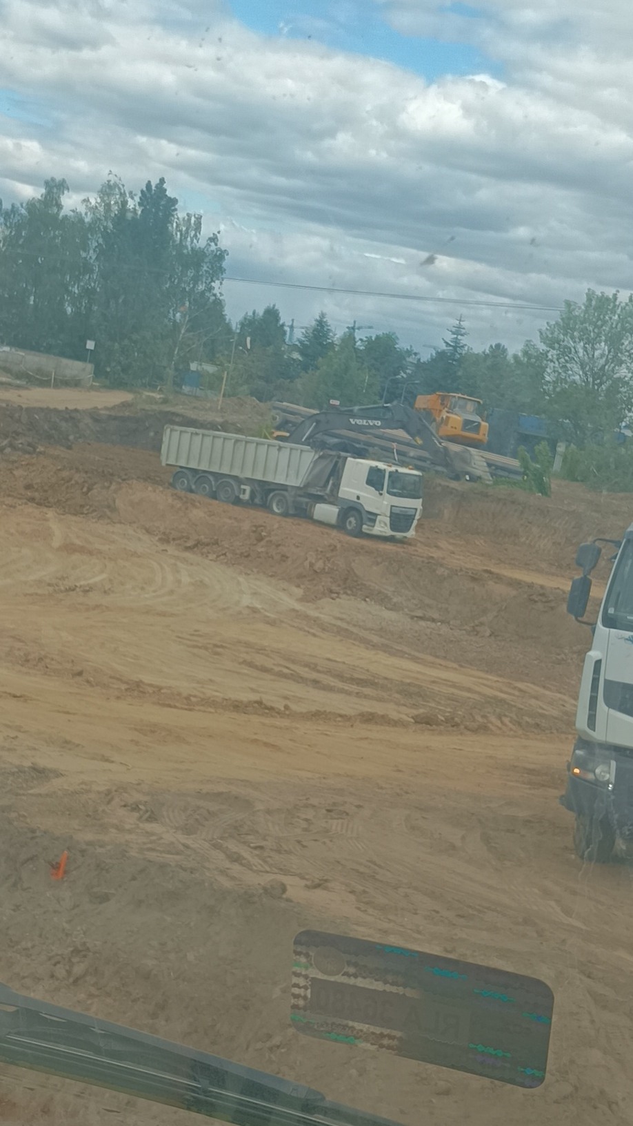 Ciężarówka wywrotka na placu budowy z koparką Volvo w tle. Widok z kabiny innego pojazdu, z pomarańczowym pachołkiem w lewym dolnym rogu.