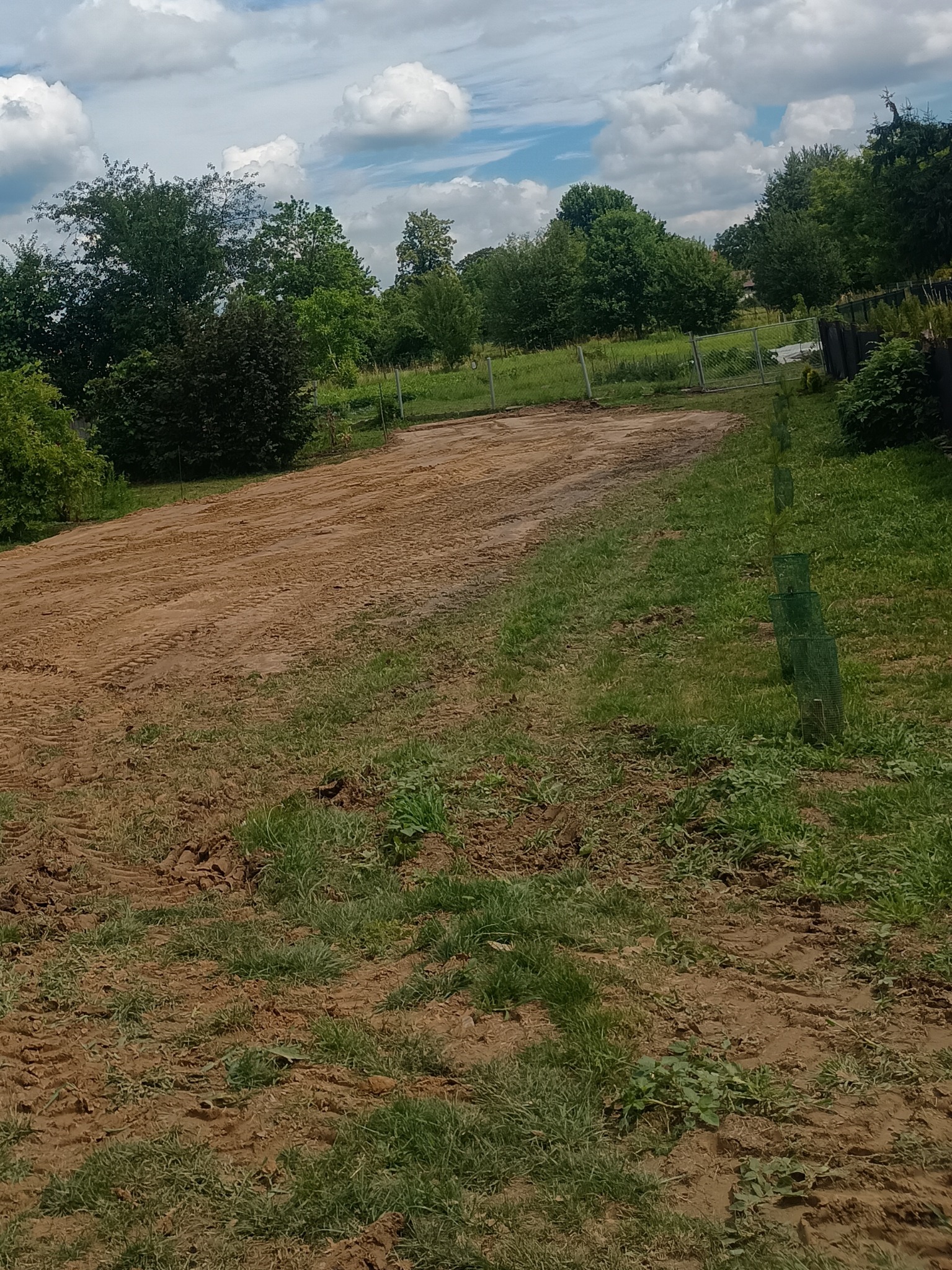 Przygotowany teren pod budowę z widocznymi śladami opon, otoczony zielenią i ogrodzeniem. Niebo z chmurami w tle.