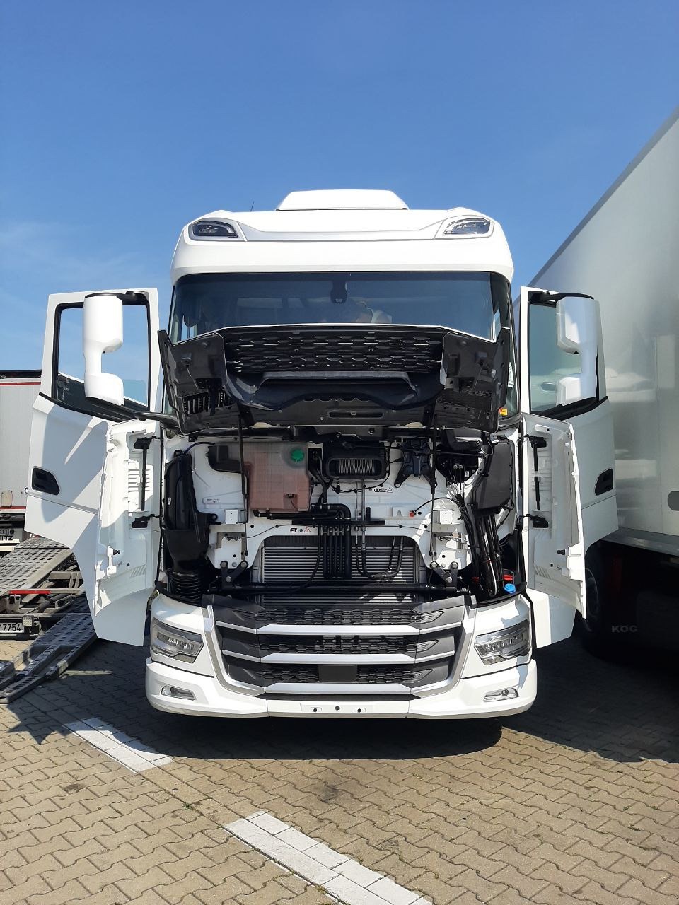 Biała ciężarówka z otwartą maską, widok z przodu na silnik i chłodnicę, zaparkowana na kostce brukowej. Widoczne otwarte drzwi kabiny i fragment naczepy.