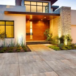 Nowoczesny podjazd z beżowej kostki brukowej prowadzący do domu z drewnianymi drzwiami i kamienną fasadą, oświetlony ciepłym światłem.