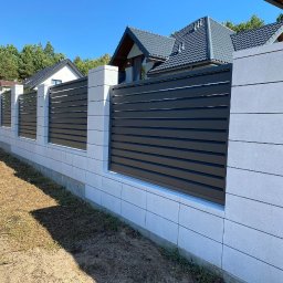 BRUKSTONE SPÓŁKA Z OGRANICZONĄ ODPOWIEDZIALNOŚCIĄ - Nowoczesne ogrodzenie panelowe z szarych prefabrykatów betonowych i poziomych, czarnych paneli, na tle domów i zieleni, w słoneczny dzień.