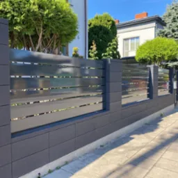 Nowoczesne ogrodzenie z szarych prefabrykatów betonowych i poziomych przęseł, na tle domu i zieleni, w słoneczny dzień.