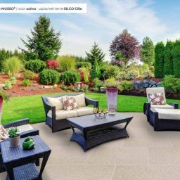 BRUKSTONE SPÓŁKA Z OGRANICZONĄ ODPOWIEDZIALNOŚCIĄ - Jasna, gładka nawierzchnia tarasu z meblami ogrodowymi z rattanu na tle zadbanego ogrodu z drzewami i krzewami. Widok na patio z kanapą i fotelem.