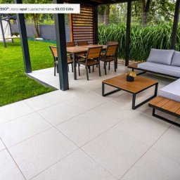 BRUKSTONE SPÓŁKA Z OGRANICZONĄ ODPOWIEDZIALNOŚCIĄ - Jasnoszara, kwadratowa kostka betonowa Musso Opal ułożona na tarasie. W tle meble ogrodowe i zieleń. Uzupełnienie SILCO Elite.