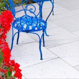 BRUKSTONE SPÓŁKA Z OGRANICZONĄ ODPOWIEDZIALNOŚCIĄ - Jasnoszara nawierzchnia z płytek tarasowych, obok błękitne meble ogrodowe i rabata z czerwonymi kwiatami. Widok z góry, delikatne światło.