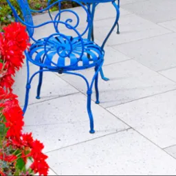 Jasnoszara nawierzchnia z płytek tarasowych, obok błękitne meble ogrodowe i rabata z czerwonymi kwiatami. Widok z góry, delikatne światło.