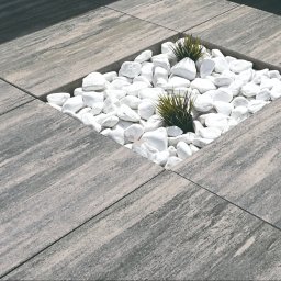 BRUKSTONE SPÓŁKA Z OGRANICZONĄ ODPOWIEDZIALNOŚCIĄ - Szare płyty chodnikowe z kwadratową wnęką wypełnioną białymi kamieniami i ozdobnymi trawami. Dekoracyjny element w przestrzeni zewnętrznej.