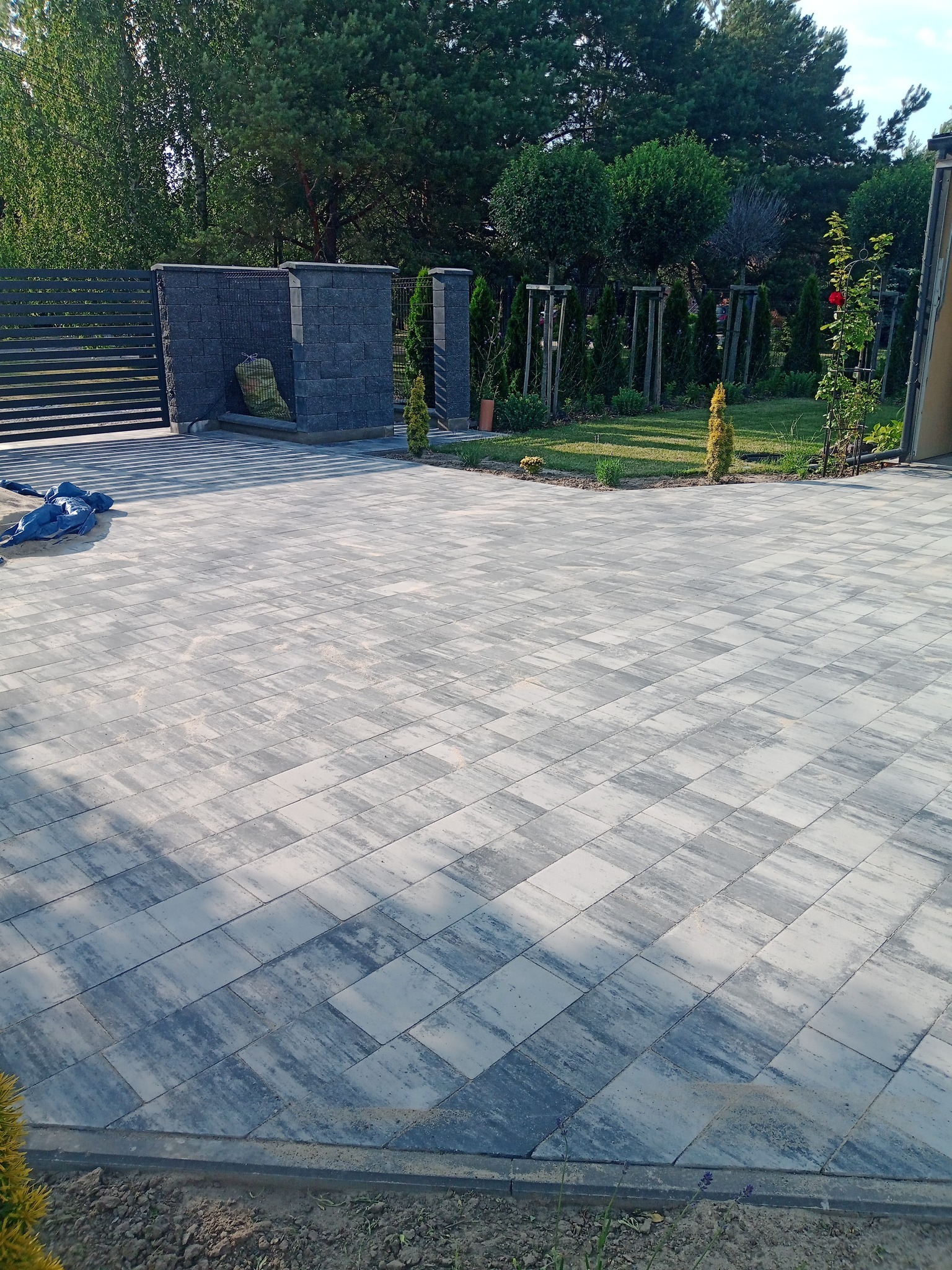 Ułożona, szara kostka brukowa tworzy wzór na podjeździe. Widoczny fragment ogrodzenia, roślinność i elementy architektury ogrodowej w tle. Słoneczny dzień.