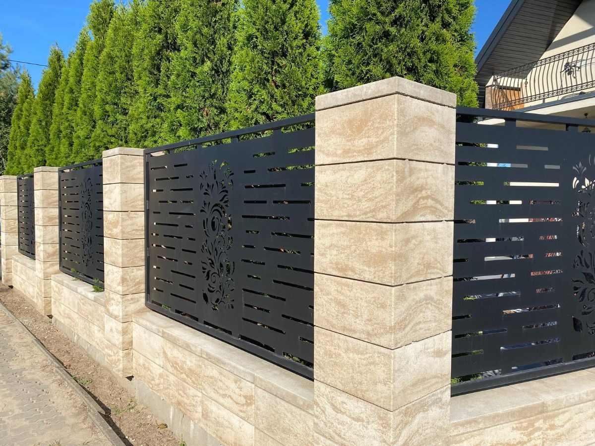 Nowoczesne ogrodzenie z beżowych bloków i czarnych paneli z motywem roślinnym, na tle zielonych drzew i domu. Układ pionowy podkreśla geometrię.