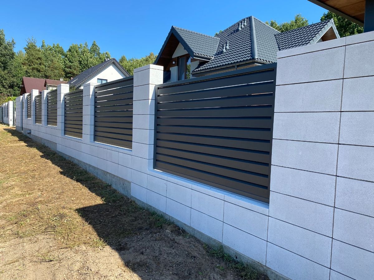 Nowoczesne ogrodzenie panelowe z szarych prefabrykatów betonowych i poziomych, czarnych paneli, na tle domów i zieleni, w słoneczny dzień.