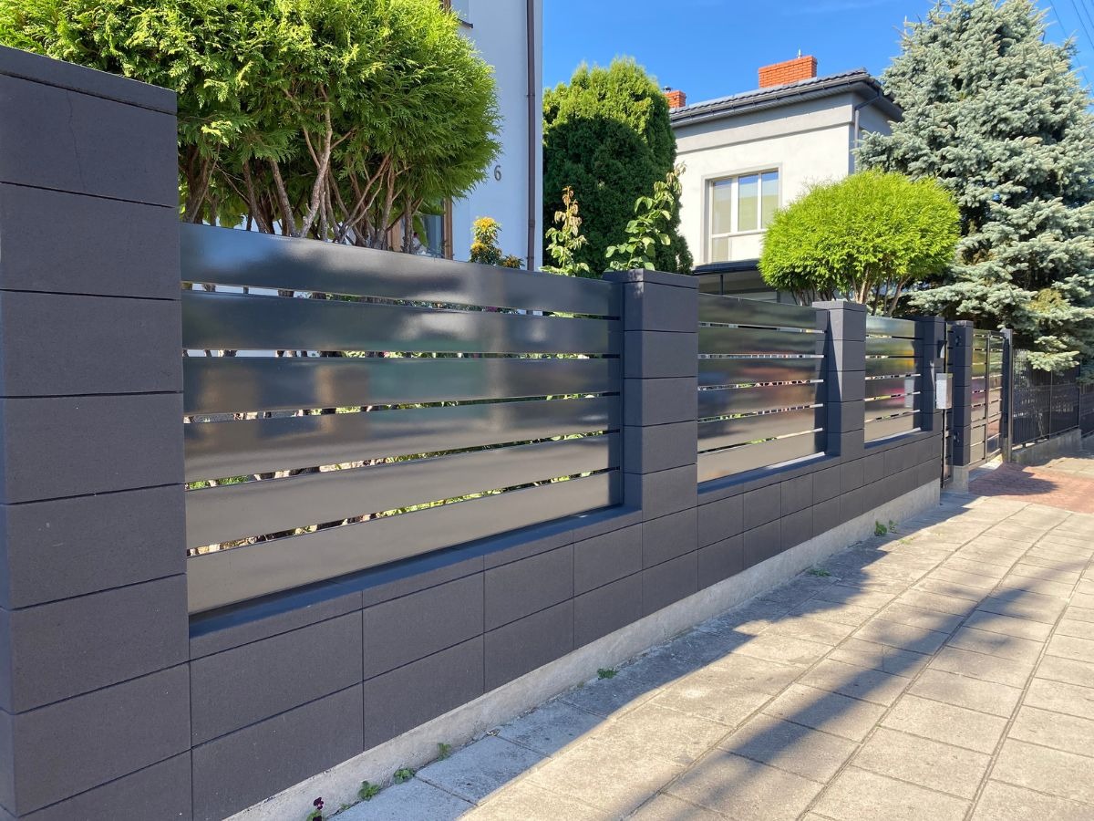 Nowoczesne ogrodzenie z szarych prefabrykatów betonowych i poziomych przęseł, na tle domu i zieleni, w słoneczny dzień.