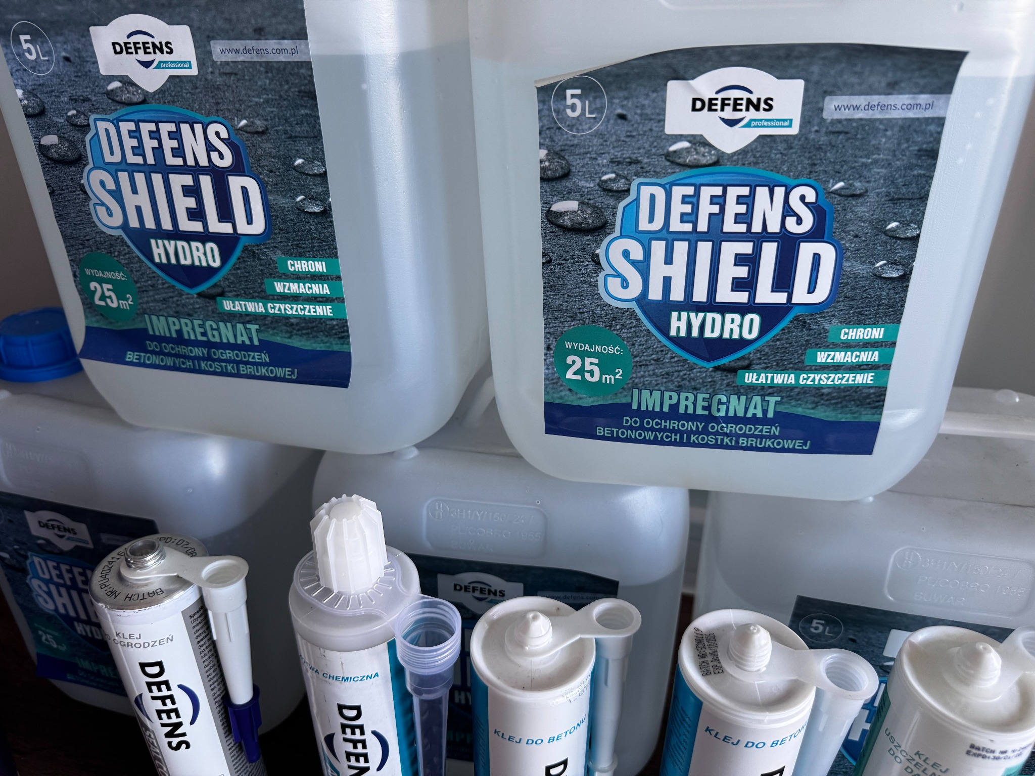 Impregnat i klej do betonu Defens Shield Hydro w białych opakowaniach, przeznaczone do ochrony ogrodzeń betonowych i kostki brukowej, widoczne etykiety z opisem produktu.