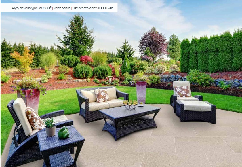 Jasna, gładka nawierzchnia tarasu z meblami ogrodowymi z rattanu na tle zadbanego ogrodu z drzewami i krzewami. Widok na patio z kanapą i fotelem.