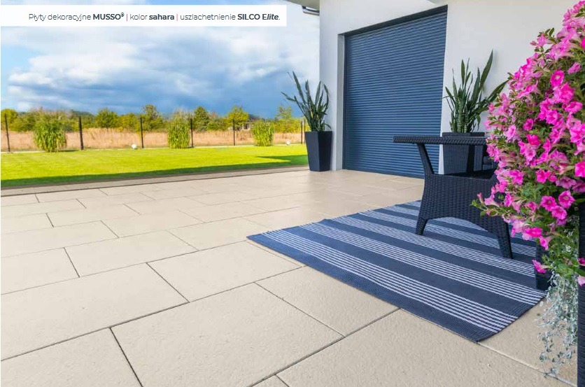 Jasna, prostokątna kostka na tarasie z meblami ogrodowymi i roślinami w donicach. W tle trawnik i błękitne rolety garażowe.