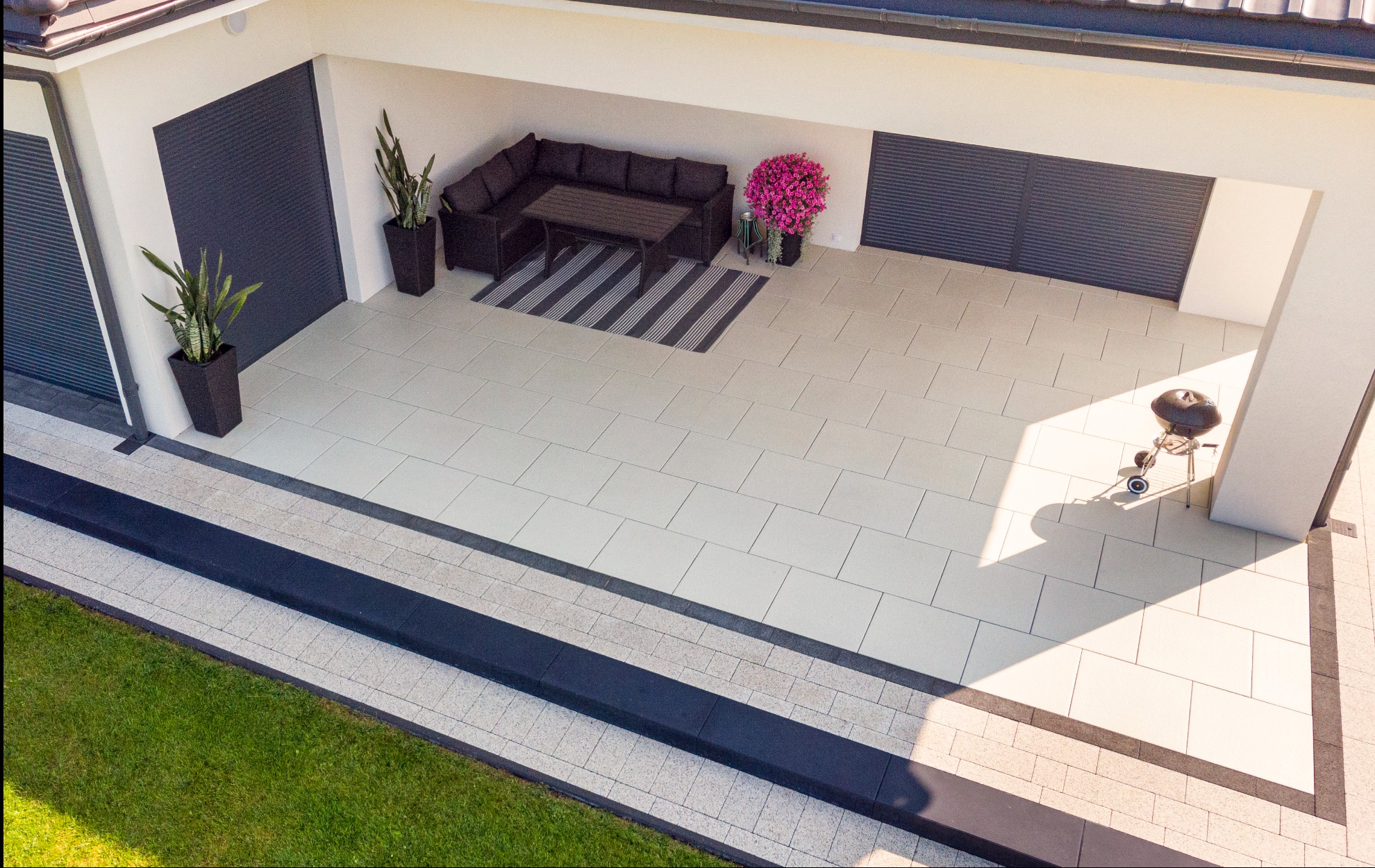 Jasna, kwadratowa nawierzchnia tarasu przylegającego do budynku, z meblami ogrodowymi, roślinami w donicach i grillem. Widok z góry.