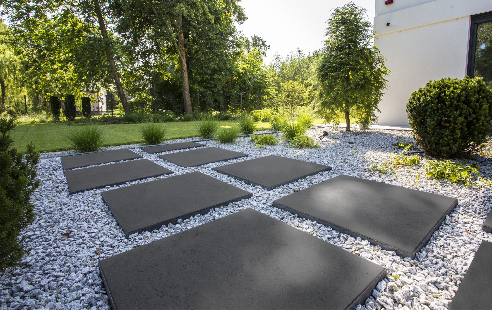 Ścieżka z płyt betonowych na białym żwirze w ogrodzie. Geometryczny wzór, nowoczesny design, zieleń drzew i krzewów w tle. Minimalistyczny styl.