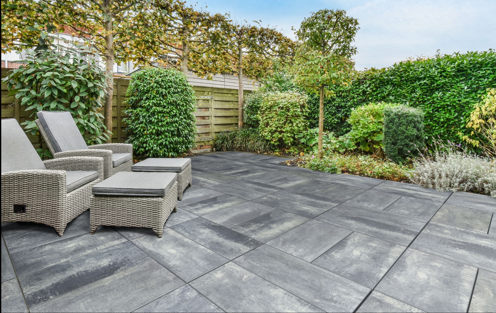 Nowoczesny taras z szarych, betonowych płyt ułożonych we wzór. W tle zielona roślinność, krzesła i pufy z rattanu.