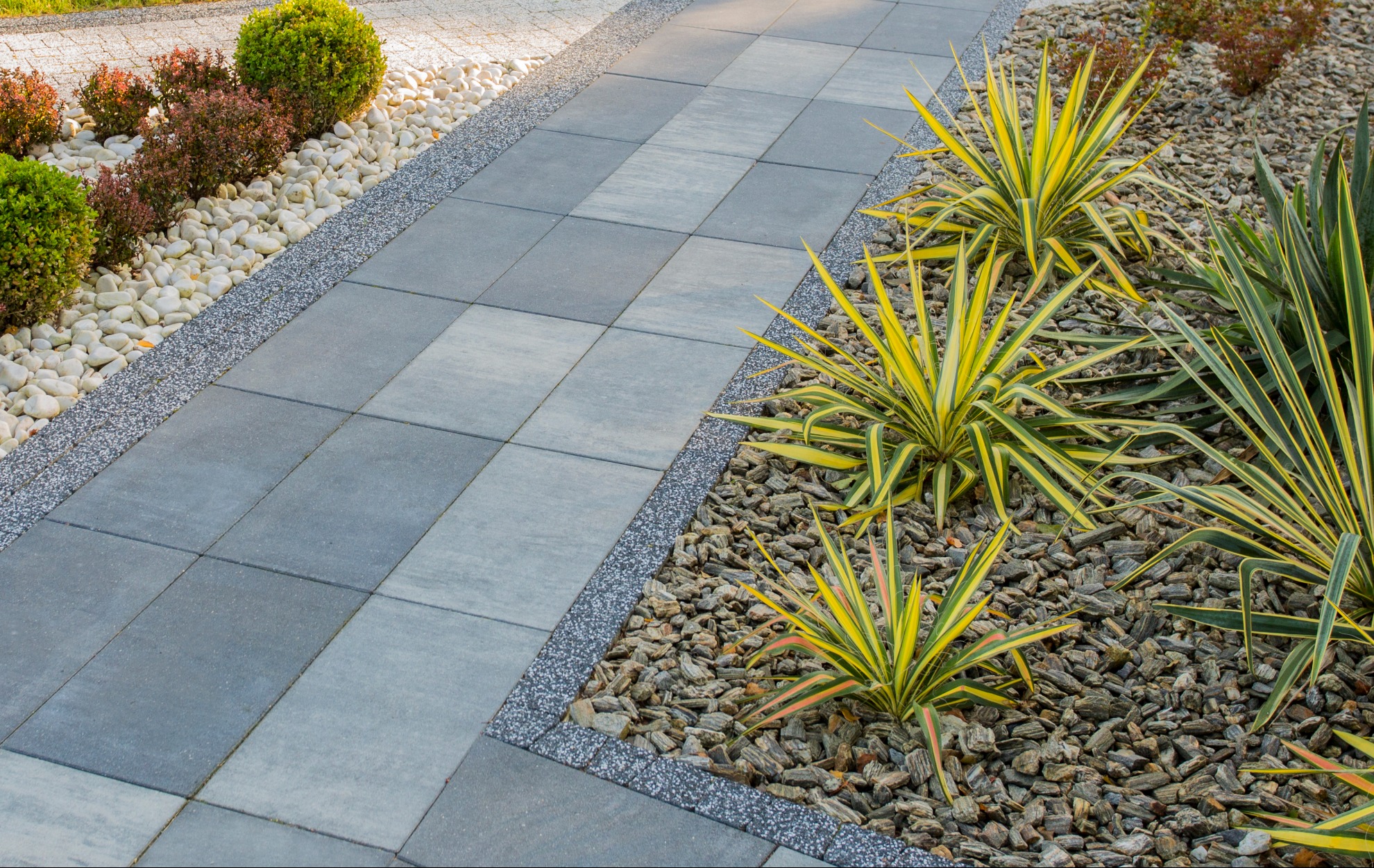 Szara, prostokątna nawierzchnia z kostki brukowej, obrzeża z drobnego żwiru, dekoracyjne kamienie i roślinność. Estetyczne wykończenie ścieżki w ogrodzie.