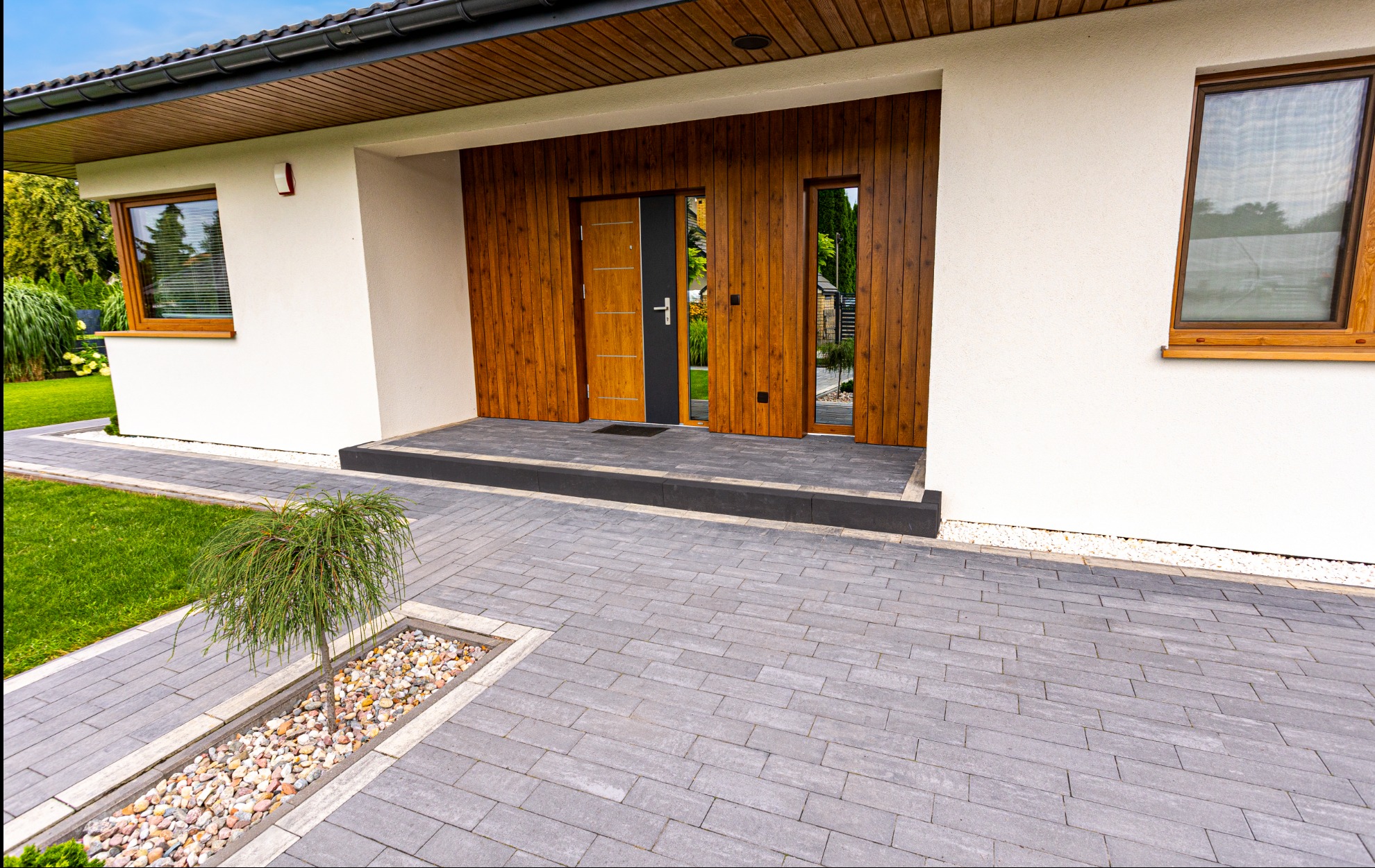 Nowoczesny podjazd z szarej kostki brukowej prowadzący do domu z drewnianymi elementami. Małe drzewko w ozdobnej rabacie dodaje uroku.