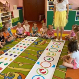 Animator w żółtej spódnicy prowadzi grę dla dzieci w sali z kolorowym dywanem i dekoracjami. Dzieci siedzą wokół mat z odciskami dłoni i stóp.