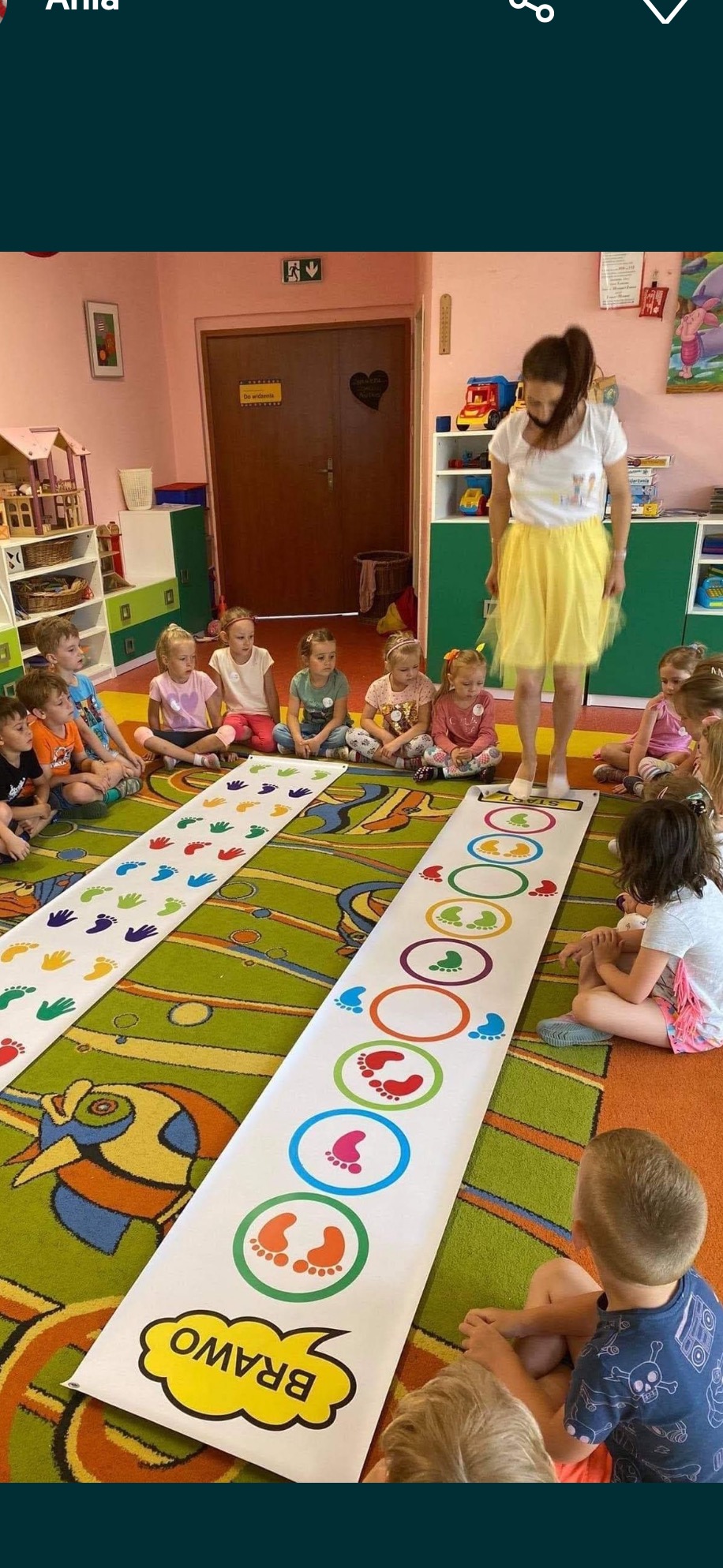 Animator w żółtej spódnicy prowadzi grę dla dzieci w sali z kolorowym dywanem i dekoracjami. Dzieci siedzą wokół mat z odciskami dłoni i stóp.