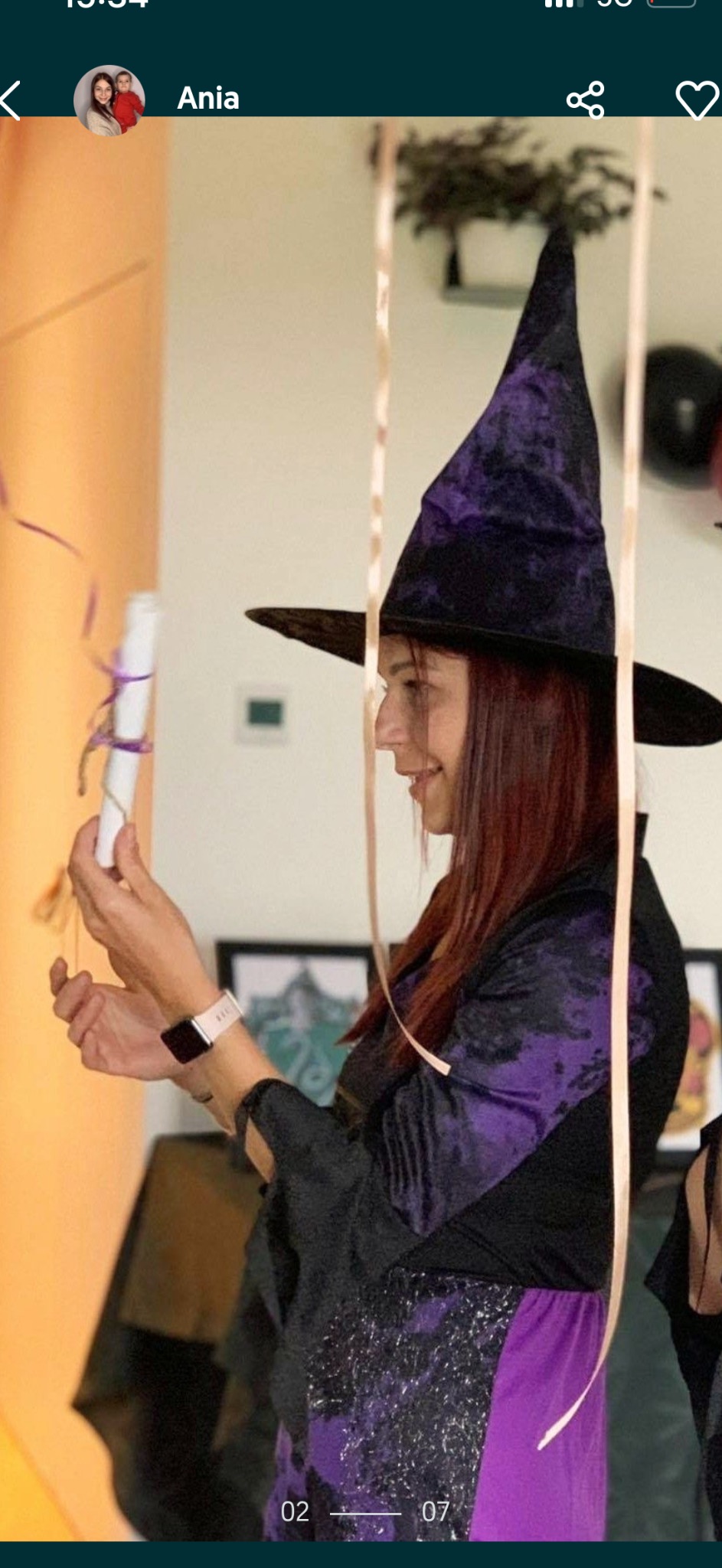 Animator w stroju czarownicy trzymająca zwój. Dekoracje urodzinowe, balony i wstążki w tle. Uśmiechnięta kobieta w fioletowo-czarnym kostiumie.