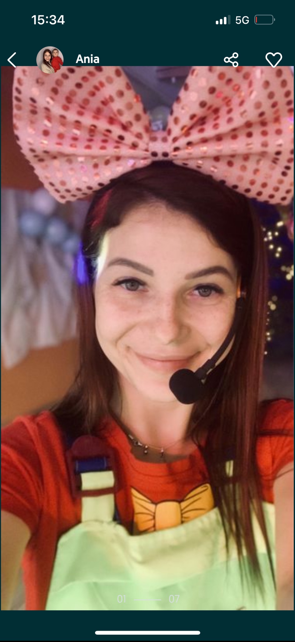 Uśmiechnięta animatorka w kostiumie z dużą różową kokardą i mikrofonem nagłownym, częściowo widoczne dekoracje imprezowe w tle.