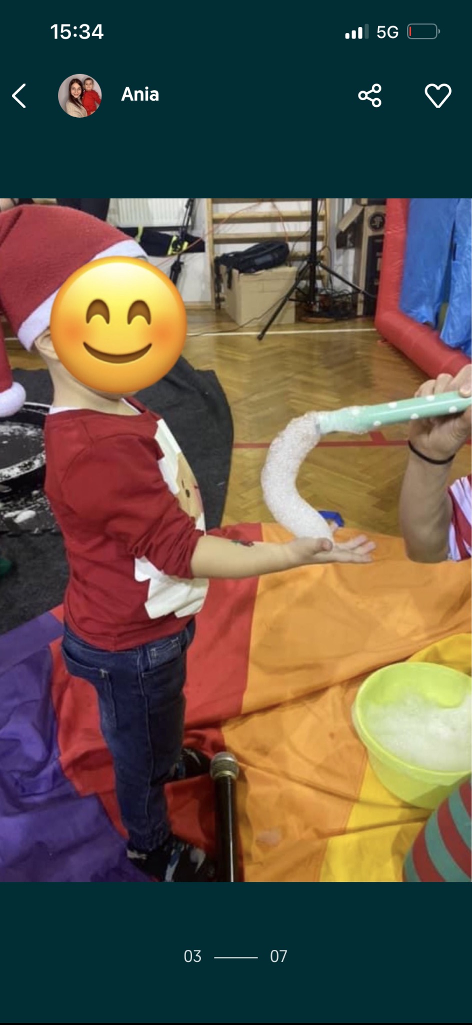 Chłopiec w czapce Mikołaja dmucha bańki mydlane z tuby na tęczowej płachcie, obok wiadro z płynem. Sala gimnastyczna w tle.